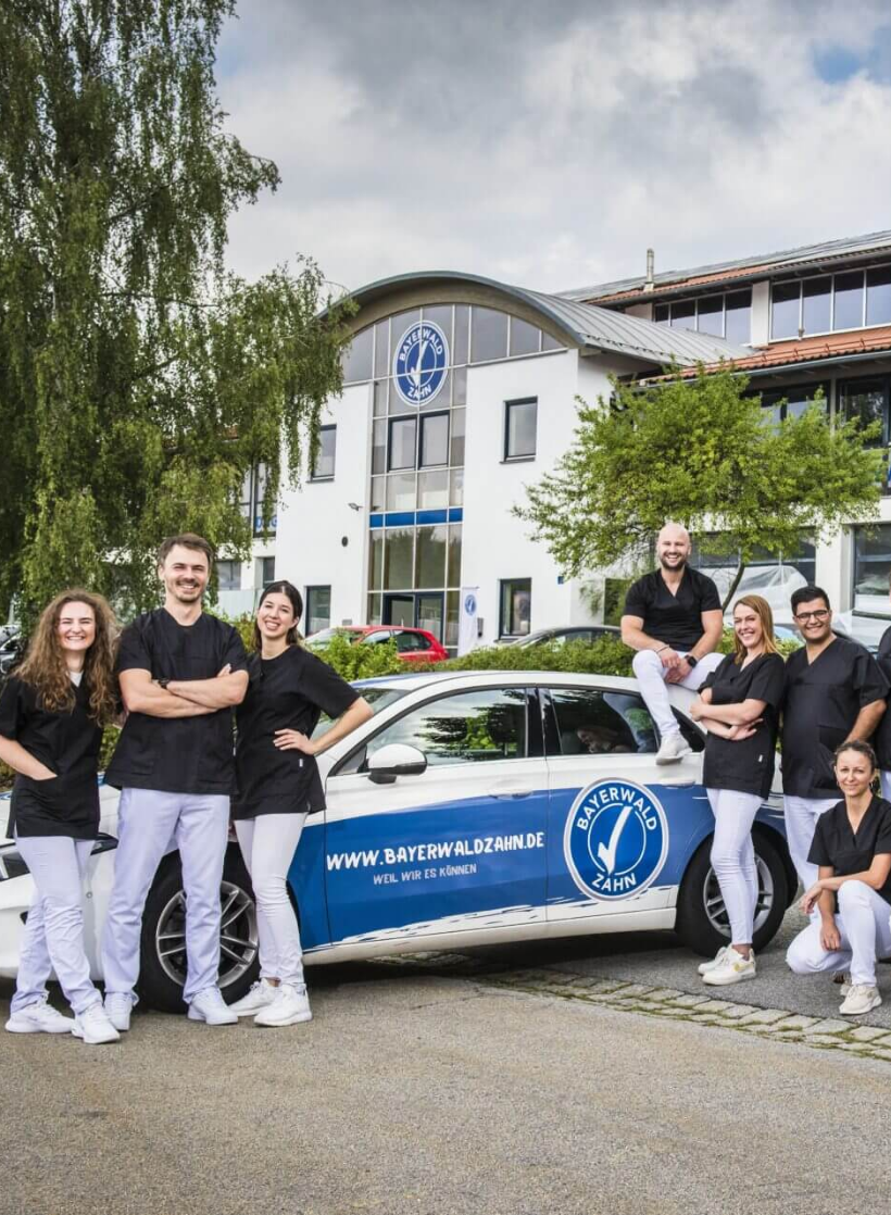 Zahnmedizinisches Team von Bayerwaldzahn steht vor Praxisgebäude neben einem gebrandeten Firmenfahrzeug.